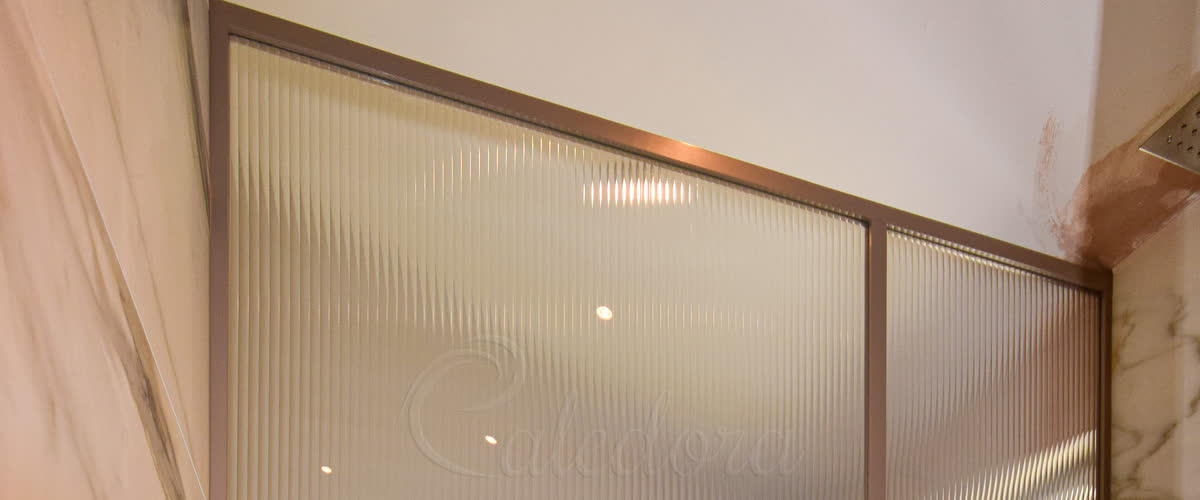 Cropped detail of a bronze-frame shower glass screen with grid-style middle bar and reeded vertical-ribbed glass in a marble bathroom
