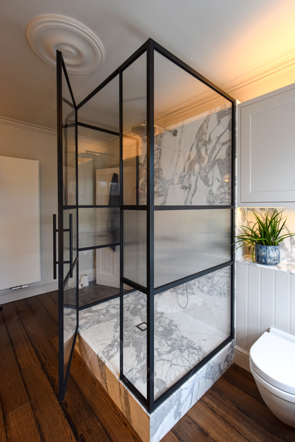 Three-sided black-frame grid glass shower enclosure with reeded lower panels and clear upper panels, hinged door open, raised marble plinth in a period London bathroom